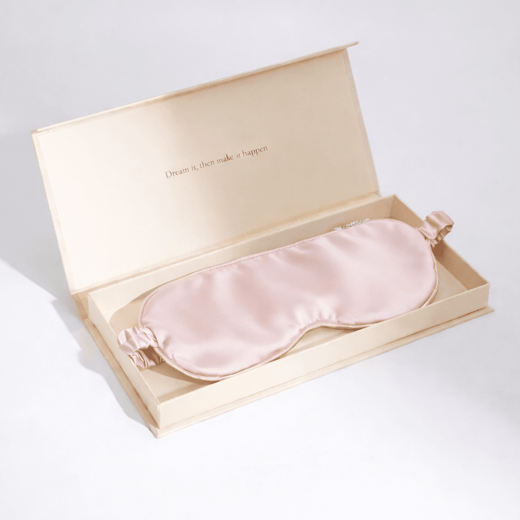 Pink silk sleep mask in an open beige box on a light grey background