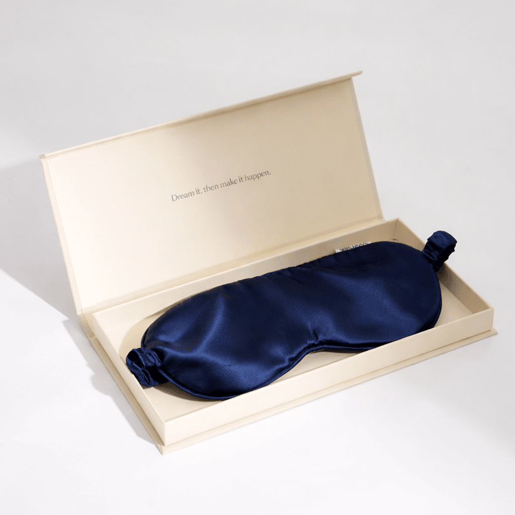 Navy blue silk sleep mask in an open beige box on a light gray background